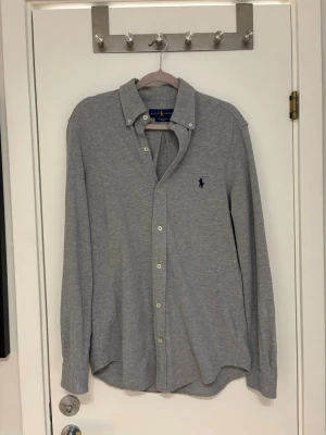 Grå skjorta från Ralph Lauren - Klassisk grå skjorta från Ralph Lauren med knappar framtill och broderad logga på bröstet. Skjortan har lång ärm, button-down krage och är tillverkad i mjuk bomull. Perfekt för en stilren och avslappnad look.
