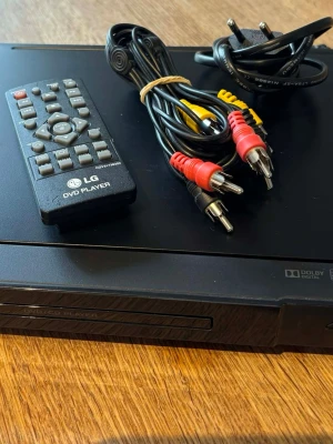 LG DVD-spelare med fjärrkontroll - Säljer en svart LG DVD-spelare med tillhörande fjärrkontroll, strömkabel och RCA-kablar (röd, vit, gul). Spelaren har en stilren rektangulär design i plast och stöd för Dolby Digital. Perfekt för att spela DVD- och CD-skivor hemma.