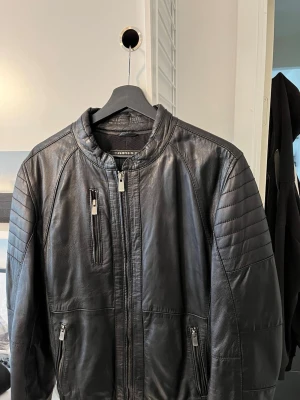 Svart skinnjacka från Jofama - Snygg svart skinnjacka från Jofama med coola detaljer på axlarna och flera dragkedjeförsedda fickor. Jackan har en modern, rak passform och står ut med sin stilrena design och höga krage. Perfekt för dig som vill ha en klassisk men trendig look. Jackan är 48 som motsvarar en M