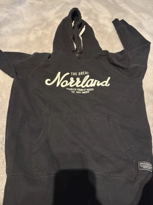 Svart hoodie The Great Norrland - Svart hoodie från The Great Norrland med vit text på bröstet och stor känguruficka framtill. Huva med vita dragsnören och ribbade muddar vid ärmslut och nederkant. Tillverkad i mjukt bomullsmaterial, perfekt för chill dagar.