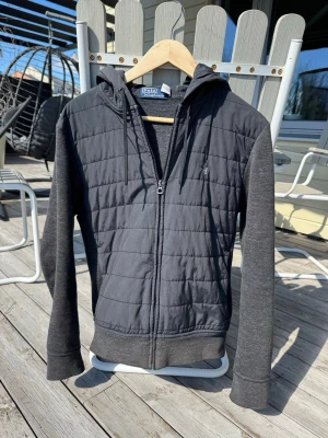 Ralph lauren cardigan  - Snygg mörkgrå cardigan i bra skick från Polo Ralph Lauren med quiltad front och huva. Dragkedja hela vägen, mjuka ärmar och rygg i sweatshirtmaterial. Bara att skriva om du har frågor!