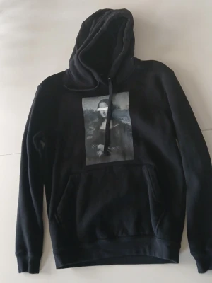 Svart hoodie med Mona Lisa-tryck - Svart hoodie från Pull&Bear med stort Mona Lisa-tryck på bröstet där ögonen är censurerade med ett vitt band. Klassisk känguruficka framtill och justerbar huva med snören. Tillverkad i mjukt bomullsmaterial, perfekt för en chill och trendig look.