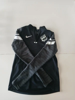Svart Nike träningströja med halv dragkedja - Svart långärmad träningströja från Nike med halv dragkedja framtill och vita detaljer på ärmarna. Tröjan har klubbmärke på bröstet och är tillverkad i ett lätt, stretchigt material som passar perfekt för sport och träning.