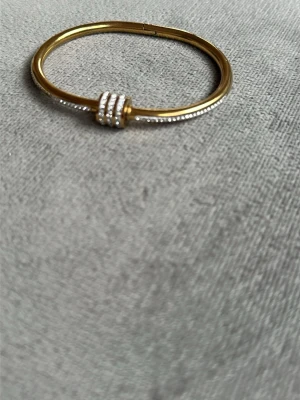 Guldigt armband med pärldetaljer - 18K guldpläterad Snyggt armband i guldfärgad metall med en rund, stel form. Armbandet har tre rader med små glittrande pärlor i mitten som ger en exklusiv känsla. Perfekt accessoar för att lyfta din outfit med lite extra bling.