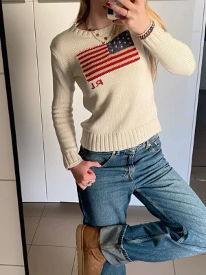  Polo Ralph Lauren flag sweter - Vit stickad tröja från Polo Ralph Lauren med amerikansk flagga och RL-broderi framtill. Tröjan har rund hals, ribbade muddar och är långärmad. Perfekt för dig som gillar klassisk trendig stil.