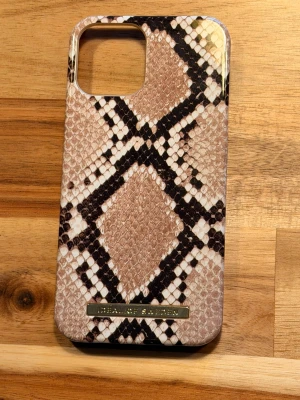 Ormmönstrat mobilskal i beige från iDeal of Sweden - Snyggt mobilskal från iDeal of Sweden med ormmönster i beige, brunt och svart. Skalet har en glansig finish och är gjort i hårdplast. Perfekt för dig som vill ge mobilen en trendig look med lite edge.