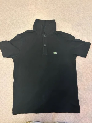 Svart pikétröja från Lacoste - Klassisk svart pikétröja från Lacoste med korta ärmar och den ikoniska gröna krokodilloggan på bröstet. Tröjan har krage och två knappar framtill. Perfekt till en avslappnad men ändå stilren look.
