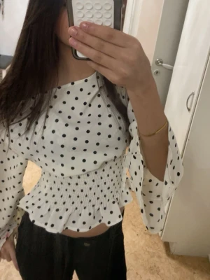Vit prickig blus med smock - Säljer en vit blus med svarta prickar och smockad midja. Blusen har långärmade, vida ärmar och en rund halsringning. Materialet känns lätt och luftigt, perfekt för att skapa en trendig look. Passar dig som gillar retrostil och att sticka ut.