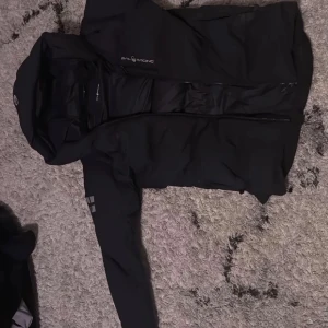 Svart parkas från Sail Racing - Svart parkasjacka från Sail Racing med huva och dragkedja framtill. Jackan har logotypdetaljer på bröstet, ärmen och huvan. Tillverkad i ett slitstarkt material som passar perfekt för kalla dagar. Snygg och funktionell design med flera fickor och justerbar huva.