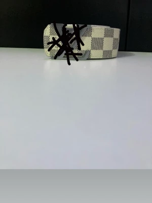 Vit och grå rutig designbälte - Snyggt bälte med vit och grå rutig design och silverfärgat spänne. Bältet har en bred form och känns riktigt trendigt, perfekt för att lyfta vilken outfit som helst. Passar dig som gillar accessoarer med lite extra attityd.