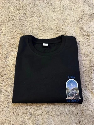  Casa Blanca t-shirt  - Svart t-shirt från Casa Blanca med ett coolt tryck på bröstet som visar en scen med palmer och byggnader. T-shirten har rund hals och korta ärmar, perfekt för dig som gillar stilrena plagg med en twist.