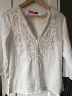 Vit broderad blus från Indiska - Luftig vit bommullsblus från Indiska med v-ringning och vackra blommiga broderier framtill. Tillverkad i ett tunt, lätt tyg som känns skönt mot huden. Den har trekvartsärm och sprund nertill i sidorna. Mycket lite använd eller inte alls.