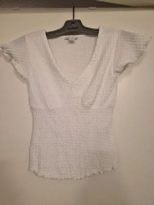 Vit v-ringad t-shirt  - Säljer en vit t-shirt från Amisu med v-ringning och korta, fladdriga ärmar. Toppen är ribbad och har en smockad struktur som ger en snygg passform. Perfekt att matcha med jeans eller kjol för en trendig look.