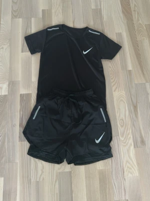 Svart träningsset från Nike - Snyggt svart träningsset från Nike med t-shirt och shorts. Båda plaggen har reflekterande detaljer och Nike-logga. Tillverkat i ett lätt och snabbtorkande material som passar perfekt för träning och sportaktiviteter.