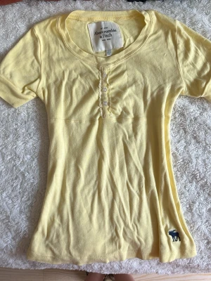 jättesöt gul tröja från Abercrombie & Fitch - Jättefin gul topp från Abercrombie & Fitch med korta ärmar, rund halsringning och dekorativa knappar framtill. Står storlek L men passar verkligen mer xs-m (har stretch så det beror på vilken fit man vill ha). Är osäker om jag vill sälja så buda gärna det ni tror är rimligt (jag säljer för billigare än köpt) då priset kan sänkas. Har inga alls synbara defekter.