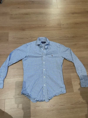 Blårutig skjorta från Ralph Lauren - Klassisk blårutig skjorta från Ralph Lauren med lång ärm och knappar framtill. Skjortan har en liten broderad logga på bröstet, Perfekt för en avslappnad men ändå stilren look.                                      Bara att höra av sig om du undrar något. Pris kan diskuteras vid snabb affär!