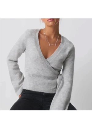 Grå omlottstickad tröja - Snygg grå stickad omlott-tröja med knytband i sidan och djup v-ringning. Tröjan har långa ärmar och är gjord i ett mjukt, fluffigt material som känns skönt mot huden. Perfekt att slänga på sig över ett linne eller t-shirt.