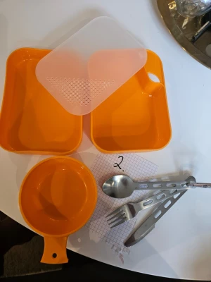Orange campingmatsats i plast - Praktisk orange campingmatsats i plast med två rektangulära matlådor, lock med sil som även fungerar som skärbräda, skål/kåsa, samt bestickset i metall (sked, gaffel, kniv). Perfekt för utflykter eller friluftsliv, allt får plats i lådan. Säljer 2 set. Detta är set 2