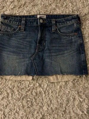 Blå jeanskjol från Monki - Säljer en blå jeanskjol från Monki med rå kant nertill och klassisk femficksdesign. Kjolen har bälteshällor och knappstängning framtill. Perfekt för dig som gillar en avslappnad och trendig stil.