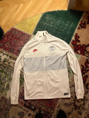 Vit Nike PSG Dri-Fit träningsjacka - Säljer en vit träningsjacka från Nike med PSG-logga och röd Nike Swoosh på bröstet. Jackan har hel dragkedja, ståkrage och två sidofickor. Tillverkad i Dri-Fit material som andas och håller dig torr under träningen. Perfekt för fotboll eller gym.