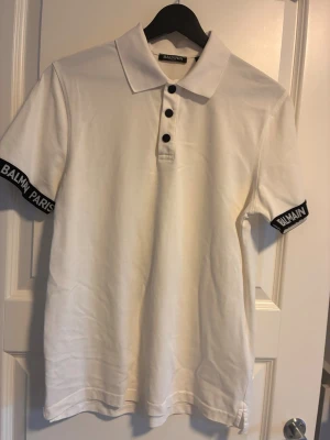 Bomull Vit piké från Balmain Paris - Säljer en clean vit pikétröja från Balmain Paris med svarta knappar och logotypdetaljer på ärmsluten. Klassisk krage och korta ärmar. Perfekt för dig som vill ha en stilren och exklusiv look.