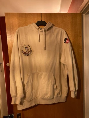 Beige Moncler hoodie med patch - Snygg beige hoodie från Moncler med stor broderad patch på bröstet där det står 'Moncler Snow Angels'. Hoodien har huva med dragsko, känguruficka och ribbade muddar. Detalj med Moncler-logga på ärmen. Perfekt för en avslappnad och trendig stil.