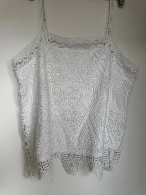 Vit spetslinne med smala axelband - Superfin vit linnetopp med tunna axelband och detaljerad spets. Toppen har ett unikt spetsmönster och dekorativ kant nertill som ger en bohemisk vibe. Perfekt att styla till jeans eller kjol för en fräsch look.