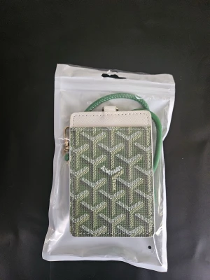 Grön korthållare med snöre - Snygg grön korthållare med klassiskt mönster och vitt parti upptill. Kommer med matchande grönt snöre och guldfärgad hake. Perfekt för att bära kort och ID runt halsen. Tillverkad i canvas och läder.