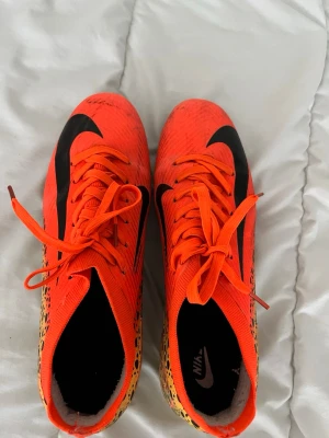 Nike Mercurial orange fotbollsskor - Säljer ett par Nike Mercurial fotbollsskor i en riktigt snygg orange färg med svarta detaljer och mönstrade sidor. Skorna har snörning och är tillverkade i syntetmaterial för bästa grepp och känsla på planen. Perfekta för dig som vill sticka ut på fotbollsplanen!