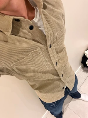 Beige overshirt i manchester - Säljer en beige overshirt i manchestertyg med svarta knappar och två stora bröstfickor. Skjortan har klassisk krage och raka ärmar, perfekt att bära öppen eller stängd över en t-shirt. Snygg och enkel att styla till jeans.