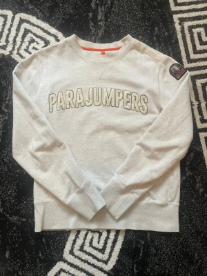 Grå sweatshirt från Parajumpers - Snygg grå sweatshirt från Parajumpers med broderad logga framtill och patch på ena ärmen. Tröjan har rund hals, ribbade muddar och en relaxed passform. Perfekt för en chill och stilren look.