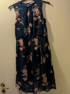 En blå klänning med blommor och fjärilar - En blå klänning med blommor och fjärilar i storlek 152 från Åhlens.                                                     Material 100% polyester, tvättas i 30 grader.                    Klänningen är längre bak än fram, en helblå underkjol och tunnare tyg med blommor/fjärilar utanpå