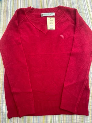 Röd cashmere tröja Abercrombie & Fitch - Snygg röd stickad tröja från Abercrombie & Fitch med V-ringning och diskret logga broderad på bröstet. Tröjan har långa ärmar och ribbade muddar vid ärmslut och nederkant. Perfekt för lager-på-lager och enkel att matcha. 100% cashmere