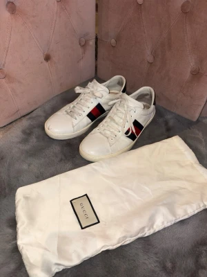 Gucci ace - Snygga vita sneakers från Gucci med klassiska röd- och blå ränder på sidan. Skorna har snörning och är tillverkade i skinn. Perfekta för dig som vill ha en stilren och lyxig look. Medföljer original dustbag från Gucci.