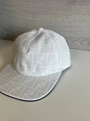 Vit keps från Fendi med FF-mönster - Snygg vit keps från Fendi med det klassiska FF-logomönstret ton-i-ton över hela kepsen. Kepsen har böjd skärm och är tillverkad i ett tygmaterial som känns lyxigt och stilrent. Perfekt accessoar för att lyfta din outfit.