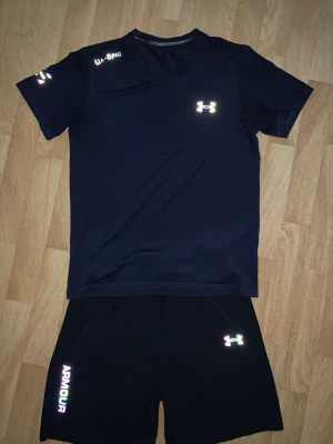 Svart träningsset från Under Armour - Säljer ett svart träningsset från Under Armour med t-shirt och shorts. Setet har reflekterande loggor och textdetaljer, perfekt för löpning eller gym. Tillverkat i ett lätt och snabbtorkande funktionsmaterial som andas.