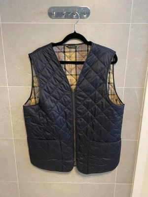 Barbour väst - Stilren marinblå quiltad väst från Barbour med klassiskt rutigt foder. Västen har två stora fickor framtill och stängs med dragkedja. Perfekt för lager-på-lager och ger en tidlös look. 