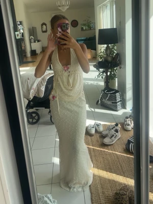Lång vit klänning med pärldetaljer - Elegant långklänning i vit nyans med pärlbroderier och blommiga detaljer. Djup v-ringning, öppen rygg och tunna axelband som knyts i nacken. Klänningen har en figurnära passform och romantiska volangdetaljer framtill.