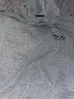 Grå Essentials Fear of God hoodie - Snygg grå hoodie från Fear of God Essentials med svart logga tryckt på bröstet och ryggen. Klassisk passform med huva och ribbade muddar. Perfekt för en avslappnad streetwear-look.