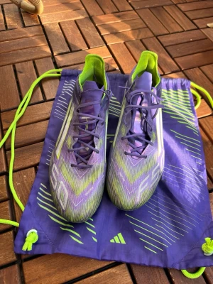 Adidas F50 lila/gröna fotbollsskor - Säljer ett par Adidas F50 fotbollsskor i lila och grönt med vita detaljer. Skorna har snörning, låg profil och är tillverkade i syntetmaterial för lätt vikt och bra bollkontroll. Medföljer matchande skopåse i samma färgtema. Perfekta för dig som vill sticka ut på planen. Storlek 46. Knappt använda. 