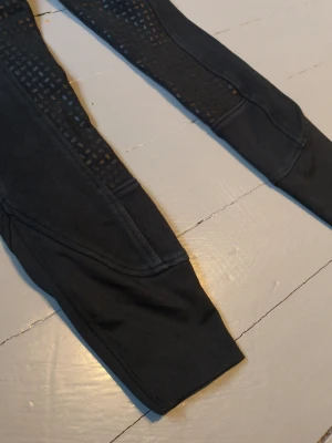 Svarta ridbyxor med silikonhelskoning - Säljer ett par svarta ridleggings med helskoning i silikon för extra grepp. De har en tight och sportig passform, stretchigt material och breda muddar vid bensluten. Perfekta för träning i stallet eller ridning. Liten defekt på baksidan. Diskret ficka framtill för småsaker.