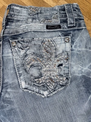 Miss Me ljusblå jeans med strass - Säljer ett par ljusblå jeans från Miss Me med snyggt slitna detaljer och en stor strassutsmyckad lilja på bakfickan. Jeansen är bootcut  och coola dekorsömmar. Perfekta för dig som gillar statement-plagg och bling! De är upp sydda så perfekt för dig som är 160 och under