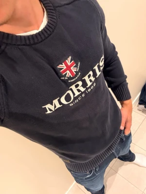 Mörkblå Morris tröja med logga - Snygg mörkblå tröja från Morris med broderad Union Jack-flagga och stor vit logotyp på bröstet. Tröjan har rund halsringning, ribbade muddar och är perfekt för en stilren och avslappnad look.