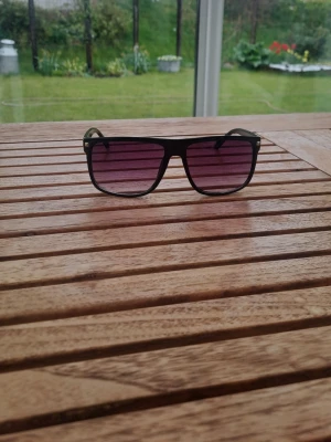 Svarta solglasögon med tonade glas till noah - Snygga solglasögon med svart båge och stora, fyrkantiga linser som är tonade i lila och rött. Perfekt accessoar för att lyfta din stil och skydda ögonen mot solen. Klassisk design som passar till det mesta. 