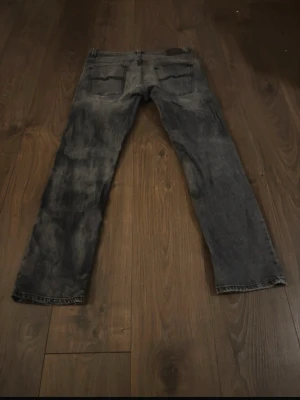 Grå jeans från Nudie Jeans - Säljer ett par grå jeans från Nudie Jeans med klassisk femficksmodell och svarta sömmar på bakfickorna. Jeansen har en rak passform och är tillverkade i slitstark bomull. Perfekta för dig som gillar en avslappnad och stilren look.