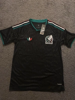 Svart Mexiko fotbollströja Adidas - Snygg svart fotbollströja från Adidas med Mexikos landslagslogga på bröstet. Tröjan har gröna och röda detaljer, klassiska Adidas-ränder på axlarna och v-ringad krage. Tillverkad i lätt och ventilerande material, perfekt för träning eller match. Storlek S men passar även i M  PRIS GÅR ATT FÖRHANDLA 