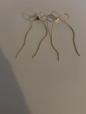Guldiga örhängen med rosettform - Säljer ett par tunna, guldiga örhängen i form av rosetter med långa, hängande kedjor. De har en minimalistisk och trendig design som passar perfekt till dig som gillar stilrena accessoarer