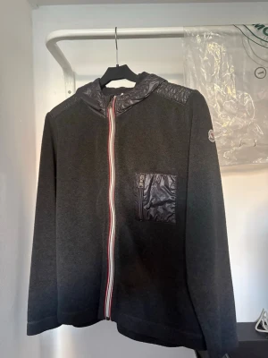 Svart hoodie från Moncler med dragkedja - Svart hoodie från Moncler med hel dragkedja framtill, huva och detaljer i glansigt material på axlar och bröstficka. Klassisk Moncler-logga på ärmen och rödvit randig dragkedja. Perfekt för en stilren och sportig look.