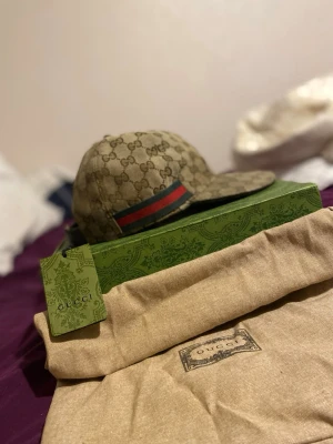 Gucci keps med GG-mönster - Snygg keps från Gucci i beige med klassiskt GG-monogram över hela kepsen. Har en grön och röd rand på sidan och justerbar rem baktill. Kepsen är tillverkad i canvasmaterial och levereras med originalpåse och låda. Priset är ej helt fast kan gå ner