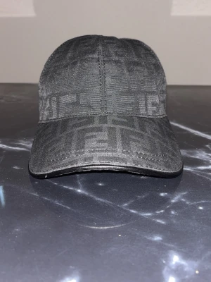 Svart keps från Fendi med monogram - Snygg svart keps från Fendi med det klassiska FF-monogrammet invävt i tyget. Kepsen har böjd skärm och justerbar baksida. Perfekt accessoar för en trendig streetwear-look. Perfekt nu för sommaren 